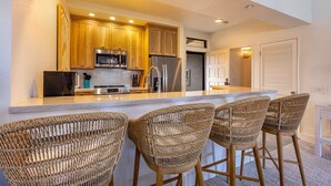 A kitchen with wooden cabinets, stainless steel appliances, and a bar area with wicker chairs. - Grand Champions Two Bedrooms - Ocean View by Coldwell Banker Island Vacations (Kihei)