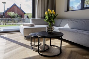 A modern living room with a grey sofa, a round black table with a vase of yellow flowers, and a view of a brick house through large windows.