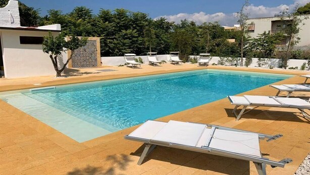 Ein rechteckiger Swimmingpool mit klarem, blauem Wasser, umgeben von einer gefliesten Liegefläche. Entlang des Poolrandes befinden sich mehrere weiße Liegestühle.