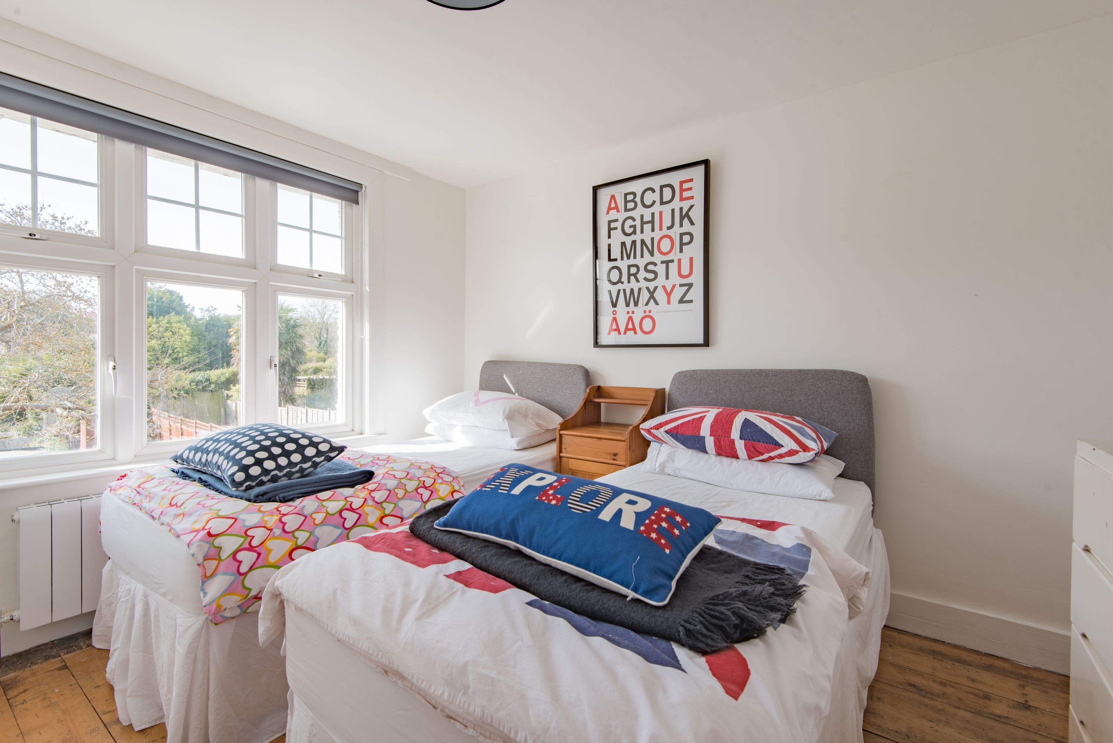 Ein Schlafzimmer mit großem Fenster, einem Bett mit vielen Kissen, einem Nachttisch und einem an der Wand hängenden Alphabet-Poster.
