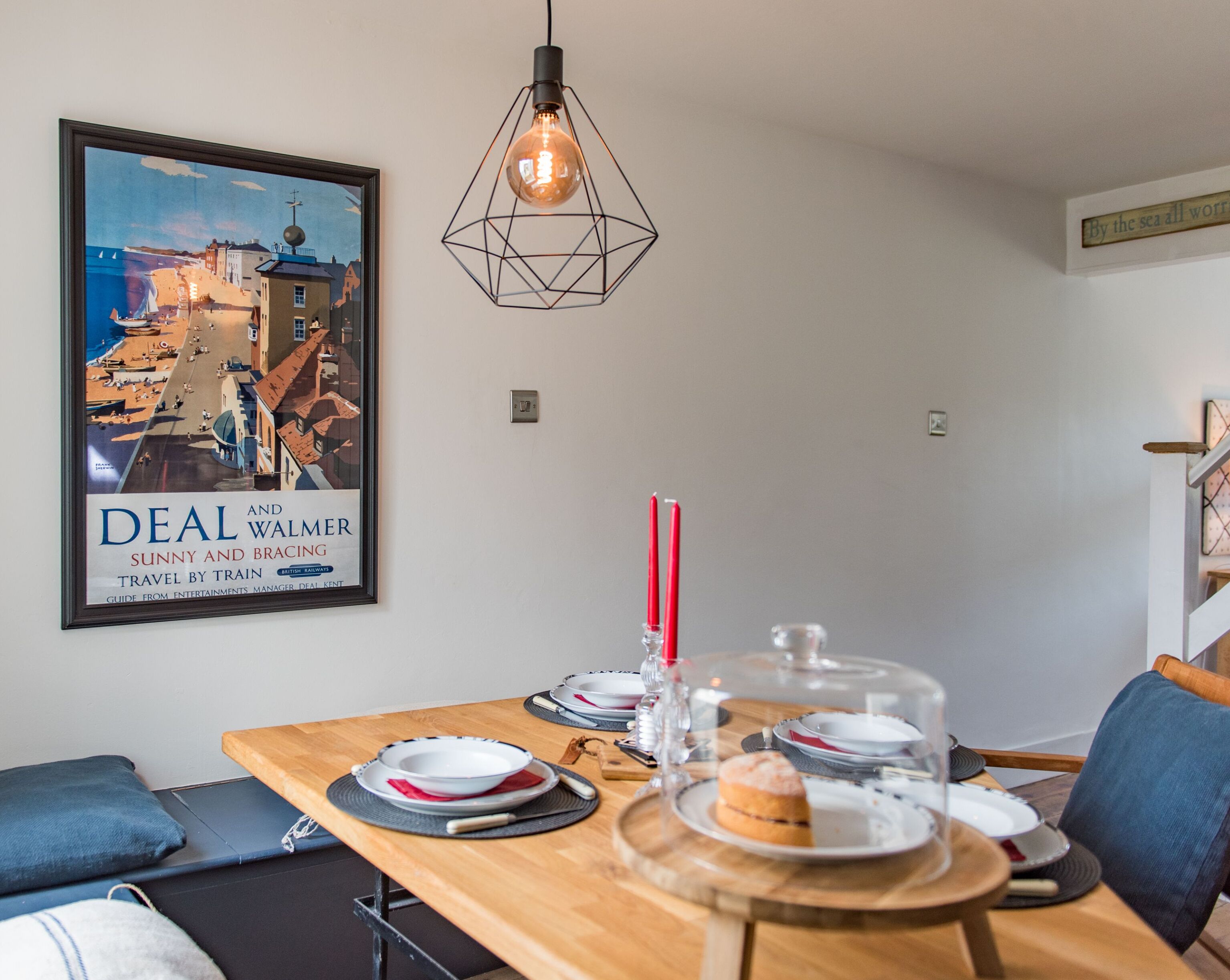 A dining area with a wooden table set for two, a framed travel poster, and a geometric pendant light.