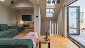 Ein modernes Wohnzimmer mit einem grünen Sofa, einem kleinen runden Tisch mit einer Flasche und Gläsern, einem Fernseher an der Wand und großen Fenstern mit Balkonblick.