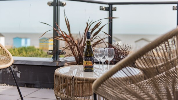Ein Korbstuhl mit einer Gavi-Weinflasche und zwei Gläsern auf einem Balkon mit Blick auf das Meer.
