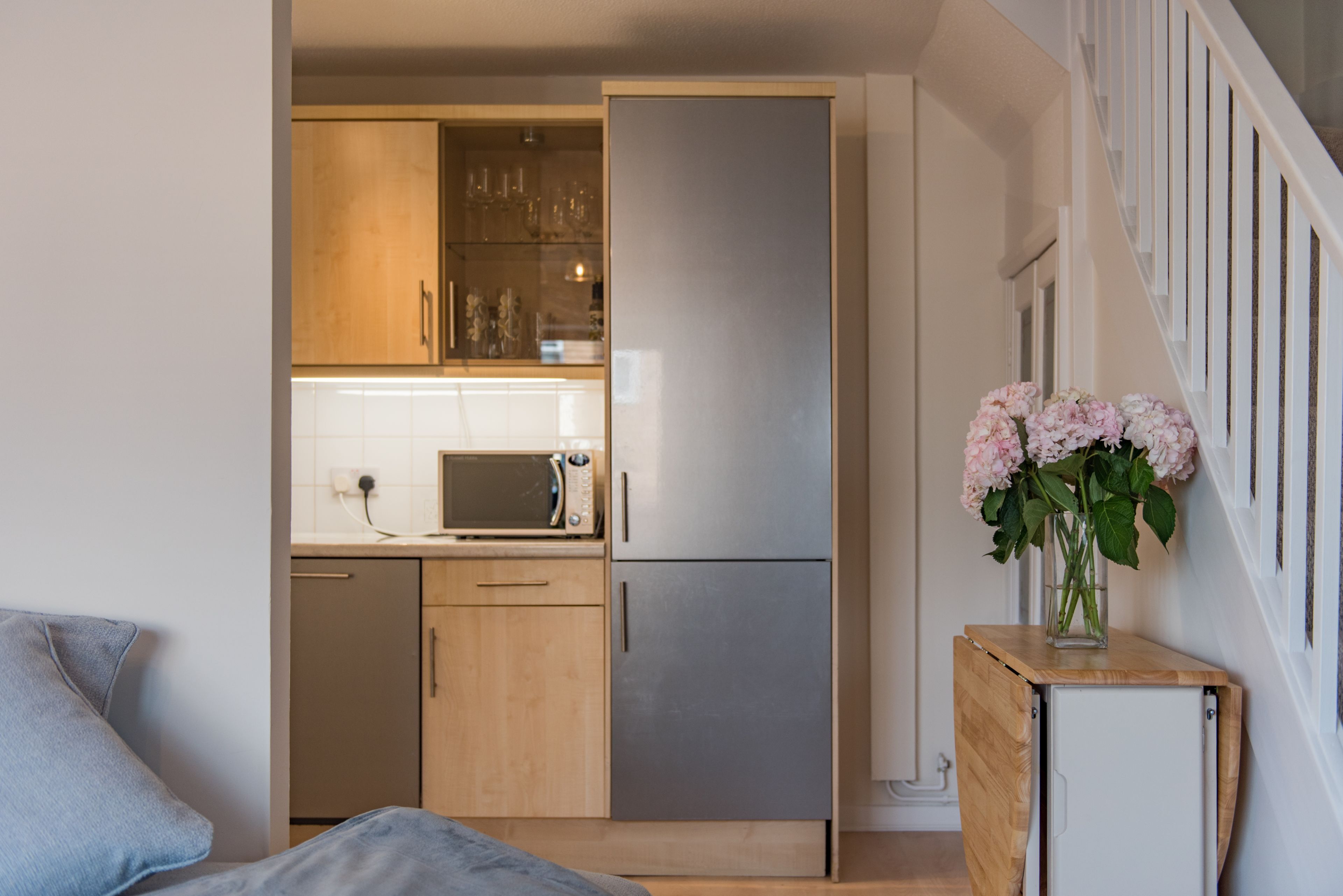 A compact kitchen with wooden cabinets, a microwave, and a refrigerator. There is a small wooden table with a vase of flowers.