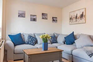 Uma sala de estar com um sofá cinzento, almofadas estampadas em azul e branco, uma mesa de centro de madeira com um vaso de flores azul, e uma parede com quatro quadros emoldurados.