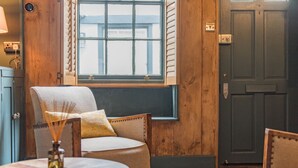 A cozy room with a wooden door, a window with shutters, a round table with a vase, and a chair with a cushion. - Charming cottage by the sea - Coop's Landing (Deal)