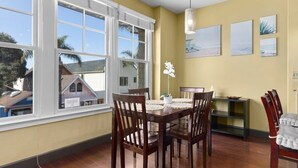 Un coin repas avec une table et des chaises en bois, un vase rempli de fleurs et une vue sur une maison, visible à travers de grandes fenêtres.