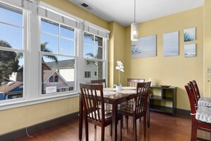 A dining area with a wooden table and chairs, large windows, and artwork on the wall.