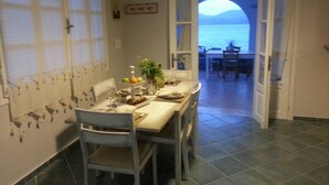 Uma área de refeições com uma mesa posta para dois, vista para o mar através de uma porta aberta e piso de azulejos.