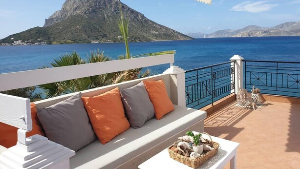 Terraço com sofá branco, almofadas laranja e uma cesta de vime com objetos decorativos. Um guarda-sol branco oferece sombra, e um corrimão preto separa o terraço do mar.