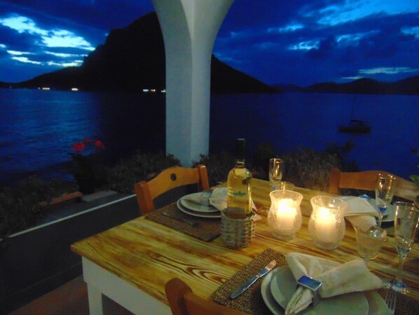 A table set for two with a bottle of wine, glasses, and a candle, overlooking a body of water at dusk.
