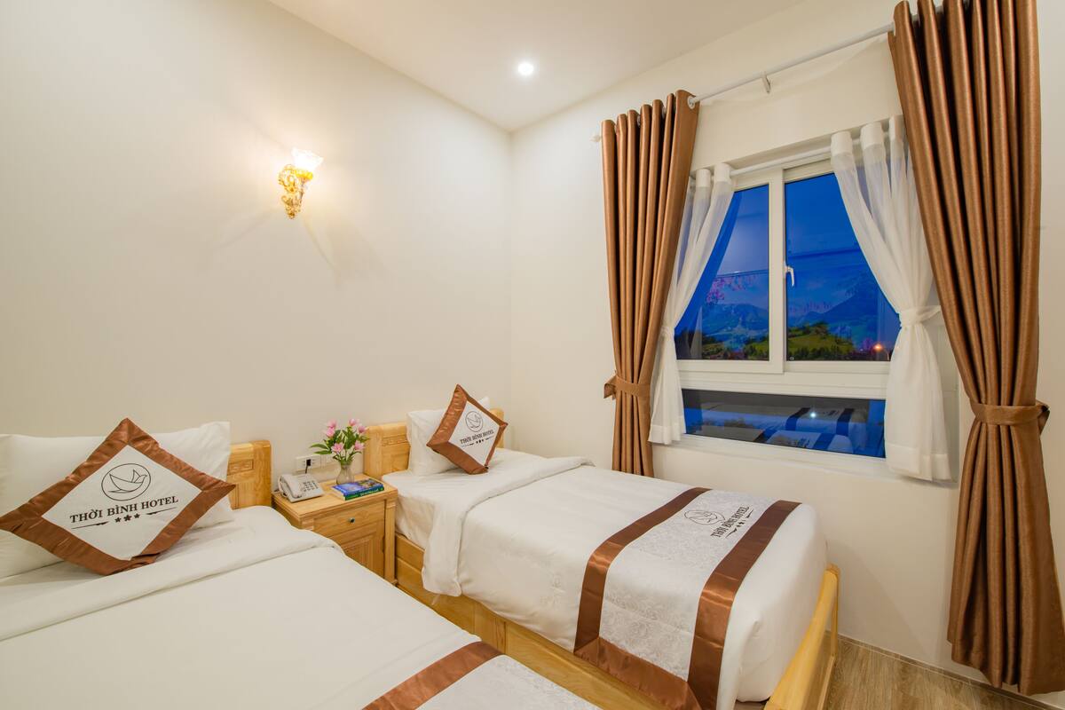 Habitación de hotel con dos camas, una mesita de noche, una lámpara y una ventana con cortinas.