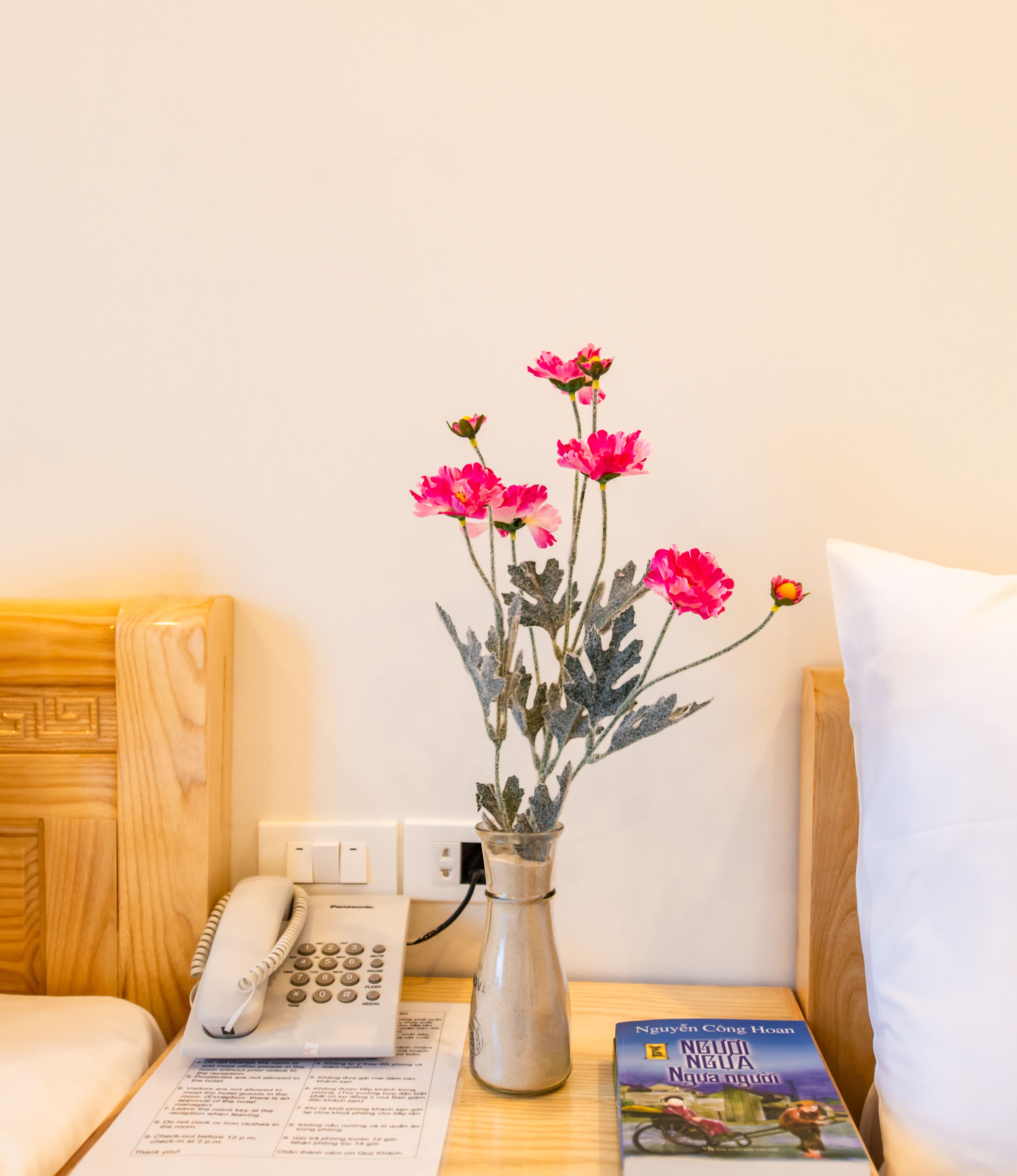 A hotel room with a wooden headboard, a bedside table with a telephone, a vase with pink flowers, and a book titled 'Nguyen Cong Huan'.