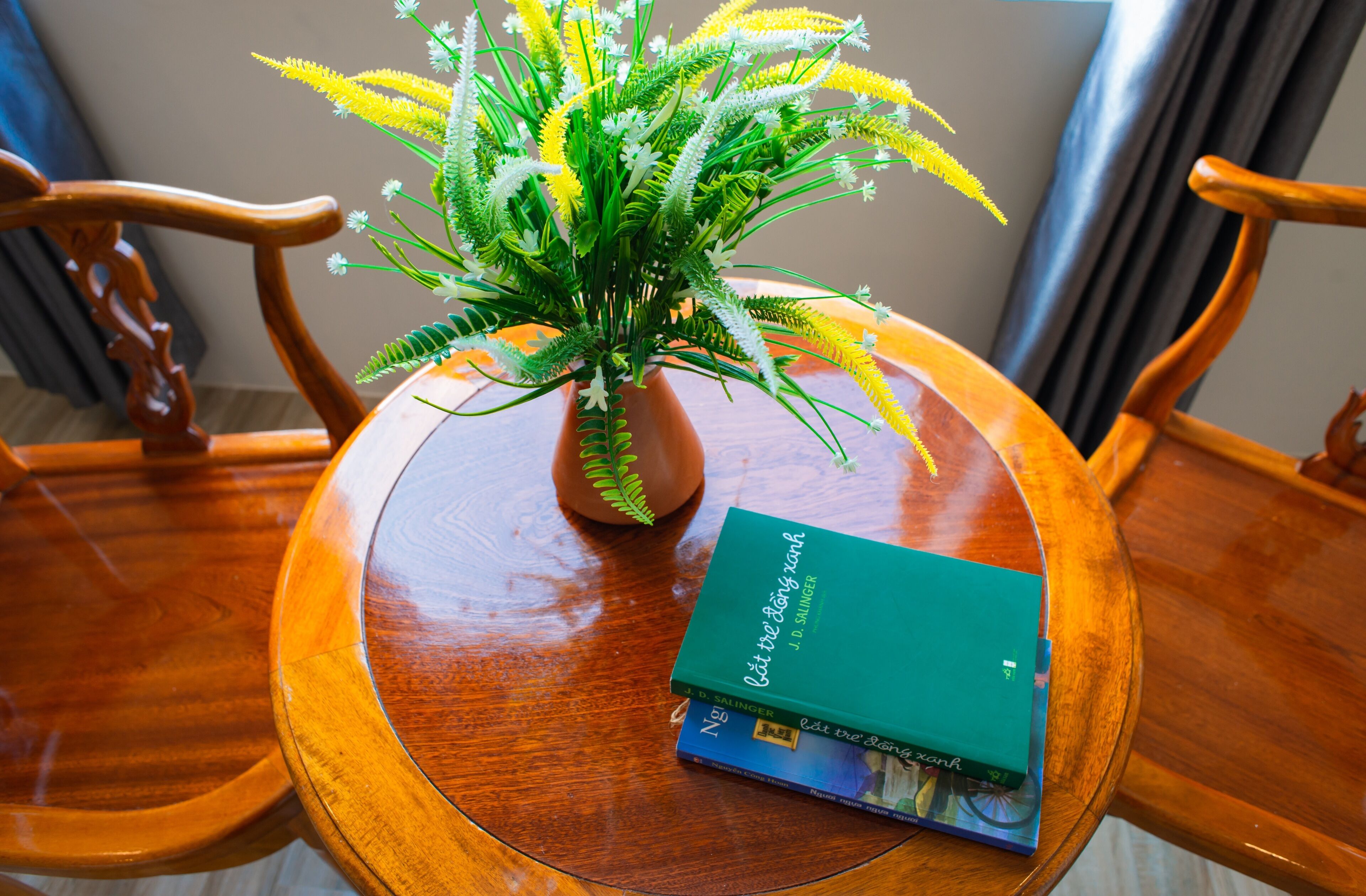 A wooden table with a green book titled "The Big Book of Singing Knots" and a potted plant with yellow and white flowers.