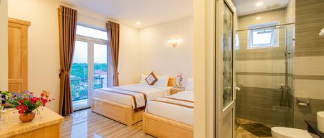 A hotel room with a wooden floor, two beds, a bathroom with a glass shower, and a view of the outdoors.