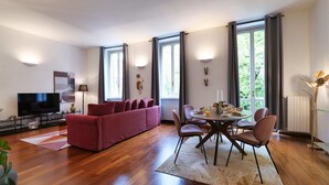 Una sala de estar moderna con un sofá rojo, una mesa de comedor con sillas, un televisor de pantalla plana y ventanas amplias con cortinas.