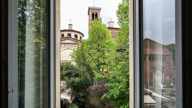 Vista a través de una ventana de un edificio con una torre del reloj y un árbol en primer plano.