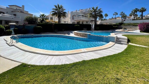A swimming pool with a curved design, surrounded by a paved deck and grass. - South facing Los Dolses bungalow - comm pool LD186 (Los Dolses)