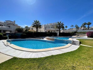 A swimming pool with a curved design, surrounded by a paved deck and grass. - South facing Los Dolses bungalow - comm pool LD186 (Los Dolses)