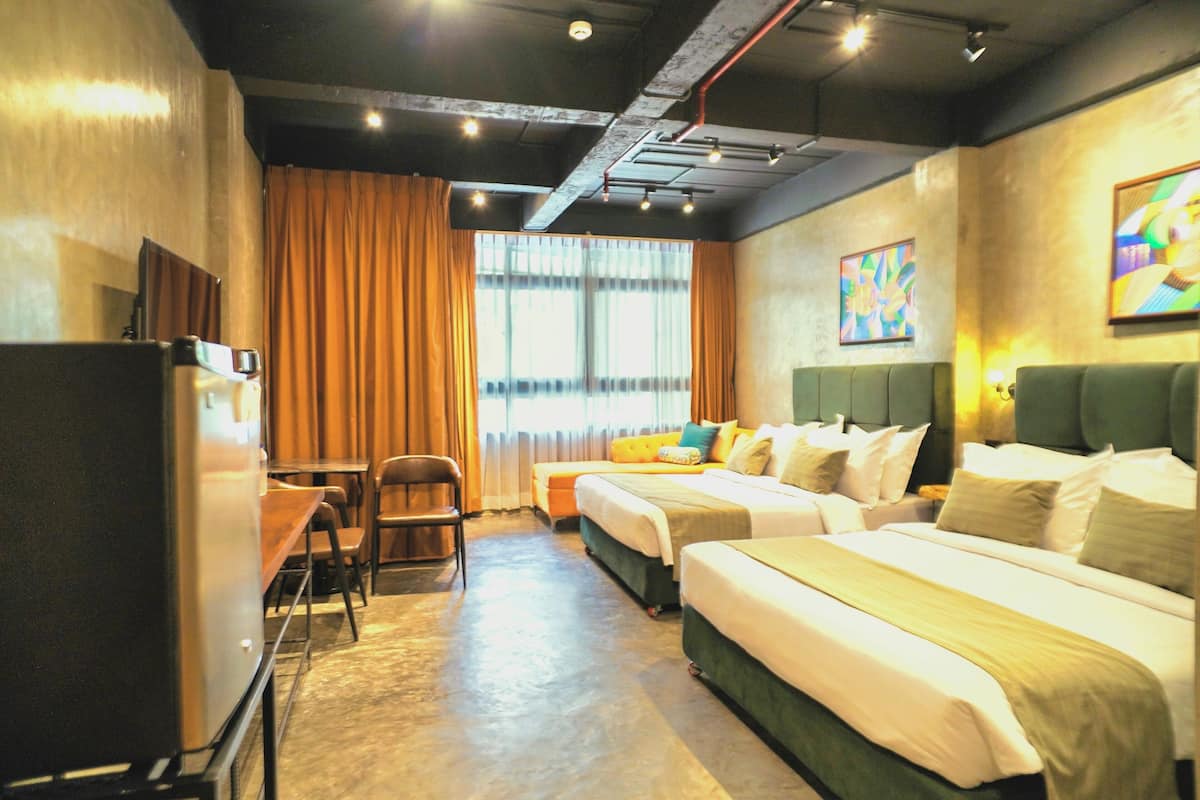A hotel room with two beds, a desk, and a chair. There is a window with curtains, a painting on the wall, and a ceiling with exposed pipes.