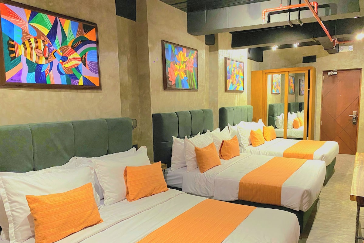 A hotel room with three beds, each with white bedding and orange accents. There are colorful paintings on the walls and a wooden door.