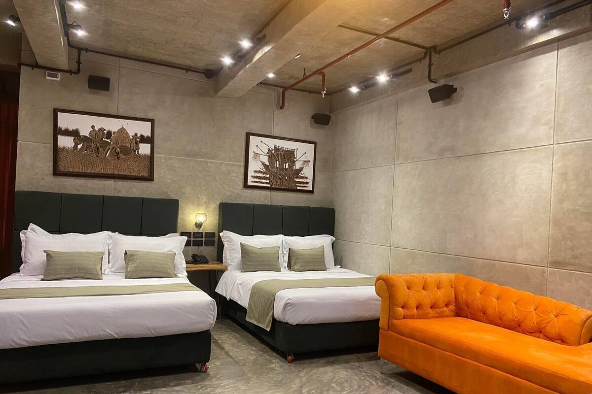 A hotel room with two beds, a nightstand, a lamp, and an orange sofa.