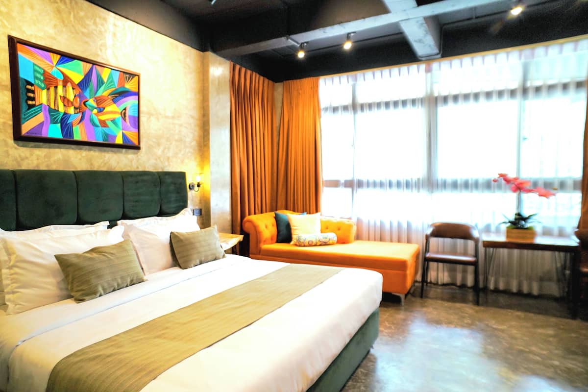 A hotel room with a large bed, a sofa, a chair, a table with a plant, and a window with curtains.