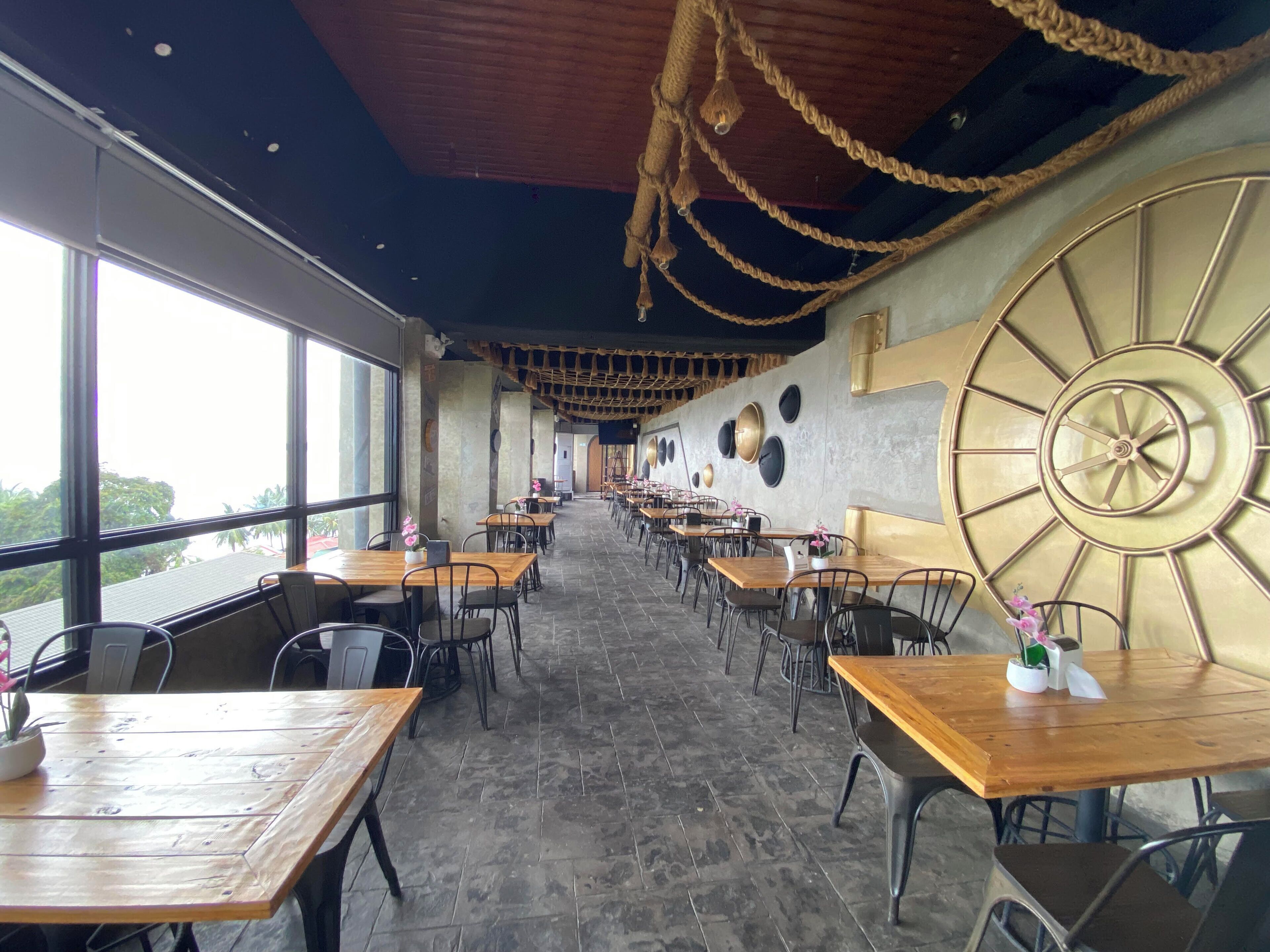 A restaurant with a long counter, wooden tables, and chairs. There are large windows on one side.