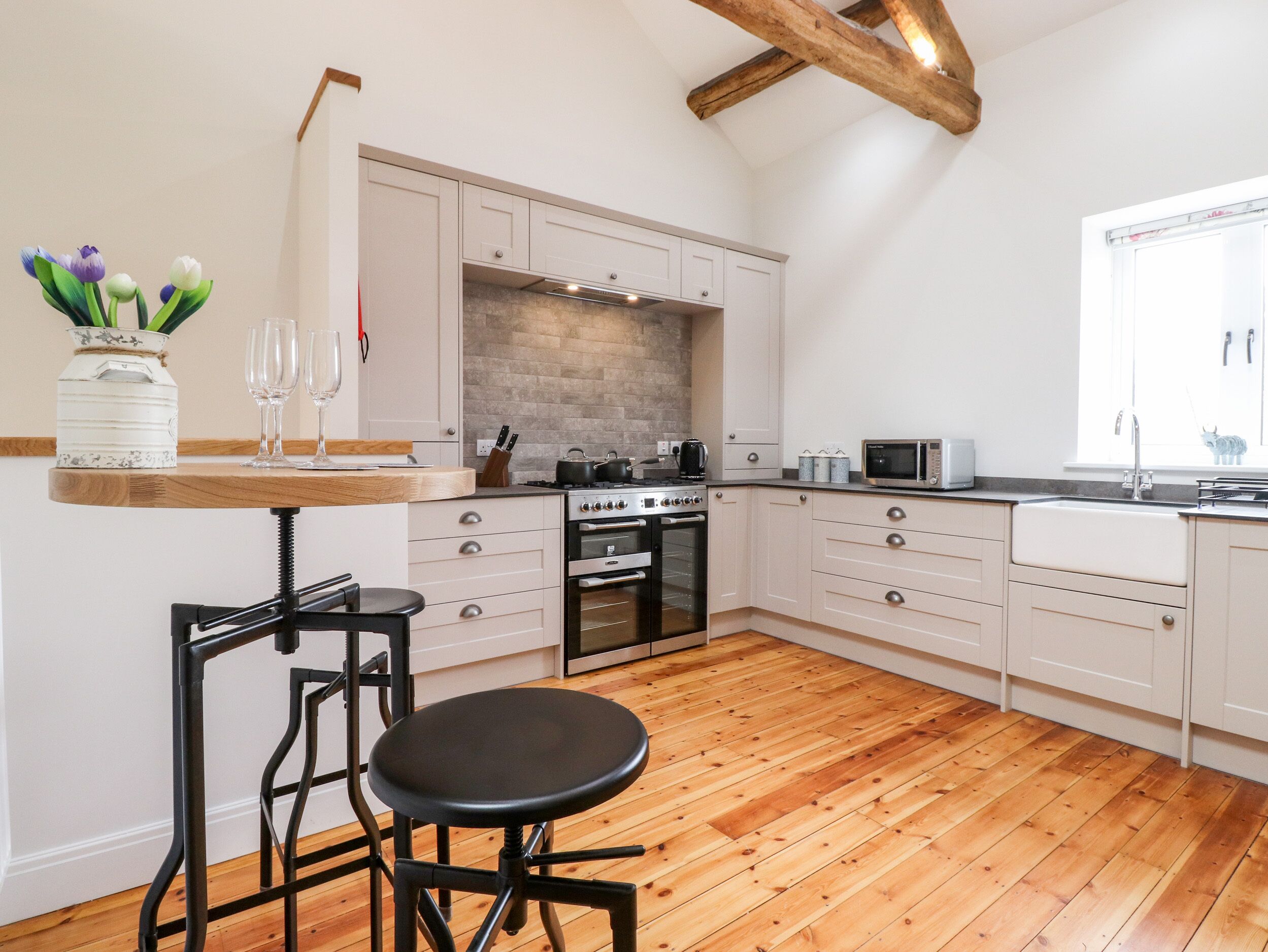 A modern kitchen with wooden flooring, white cabinets, a built-in oven, and a wooden beam ceiling.