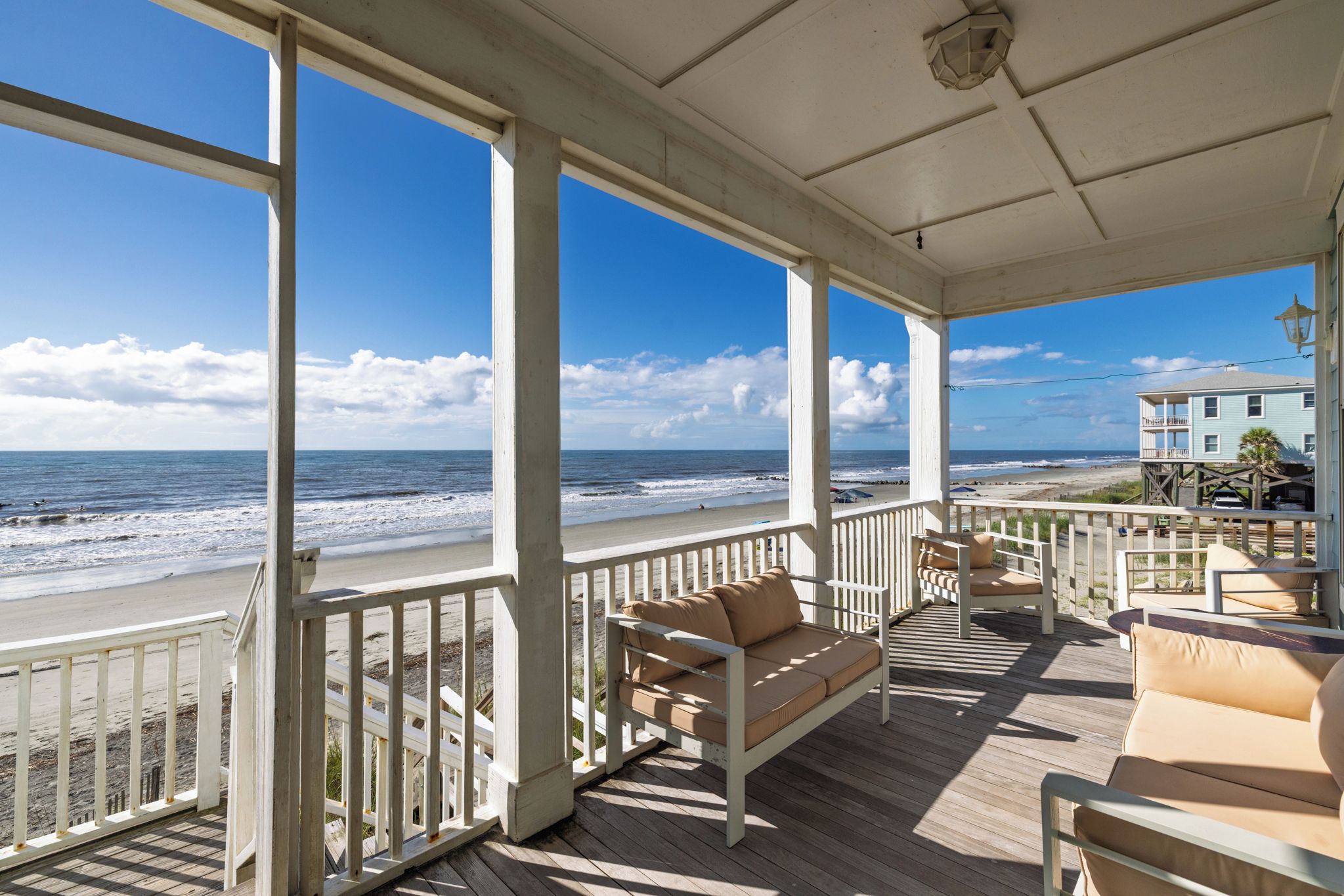 A covered outdoor deck with a view of the beach and ocean, furnished with a sofa and chairs.