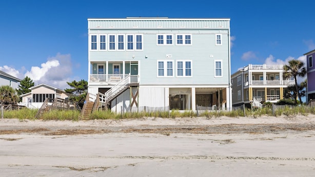 Edificio frente a la playa de dos pisos con balcón, porche cubierto y escalera que lleva a la entrada.