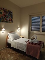 A single bed with white linens, a pink throw blanket, and a swan-shaped towel arrangement. There are two wall-mounted lights and a colorful abstract painting above the bed.