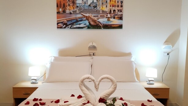 A hotel room with a bed, two bedside lamps, a painting of a cityscape, and rose petals arranged on the bed. - Palazzo Sant'Antonio Roma (Rome)
