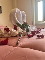 A bed with pink bedding, a heart-shaped towel, and rose petals.