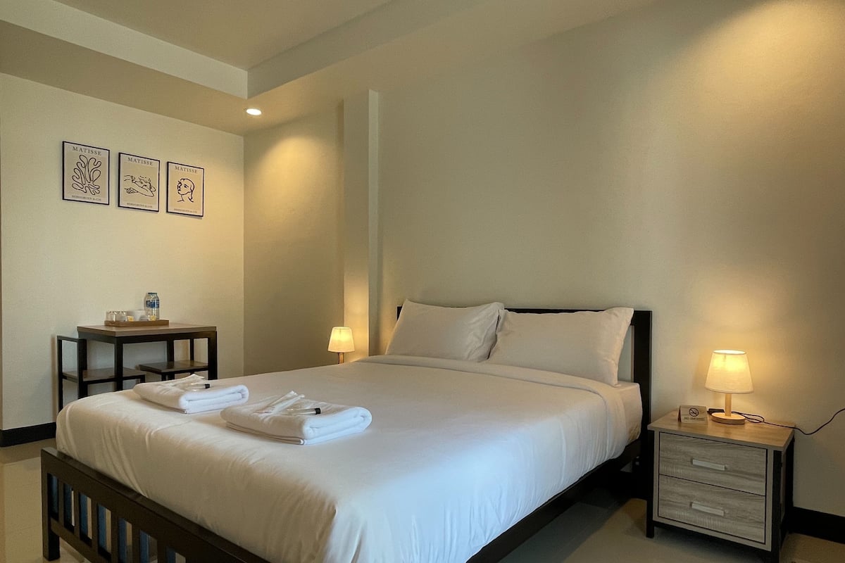 A neatly made bed with white linens and towels, a bedside table with a lamp, and wall art depicting famous artists.