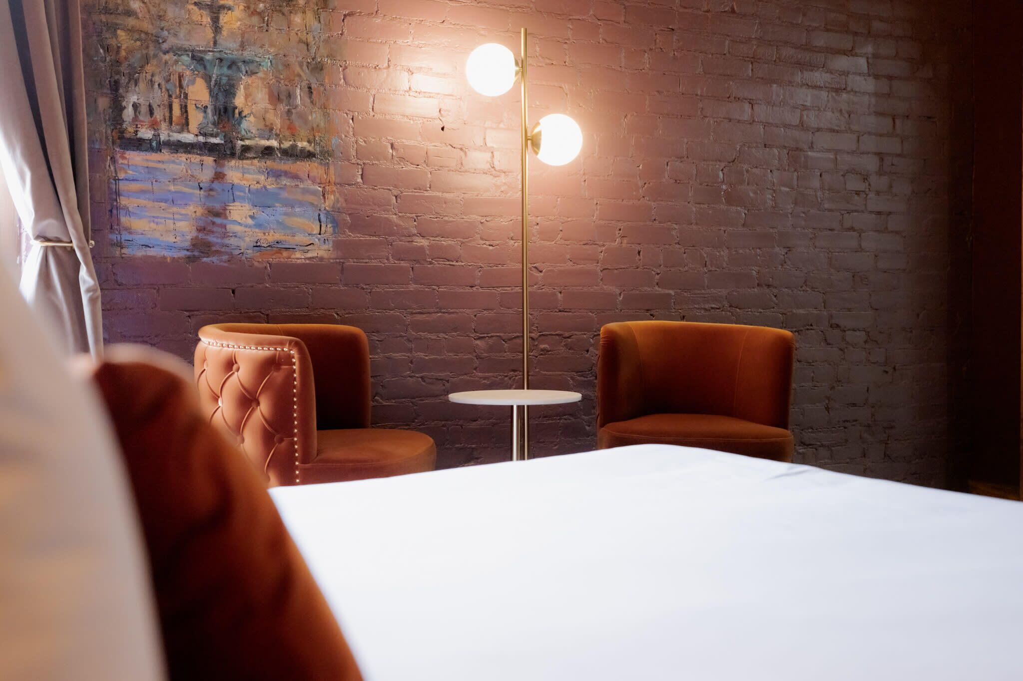 A hotel room with a brick wall, two brown leather armchairs, a small round table, and a floor lamp with two spherical lights.