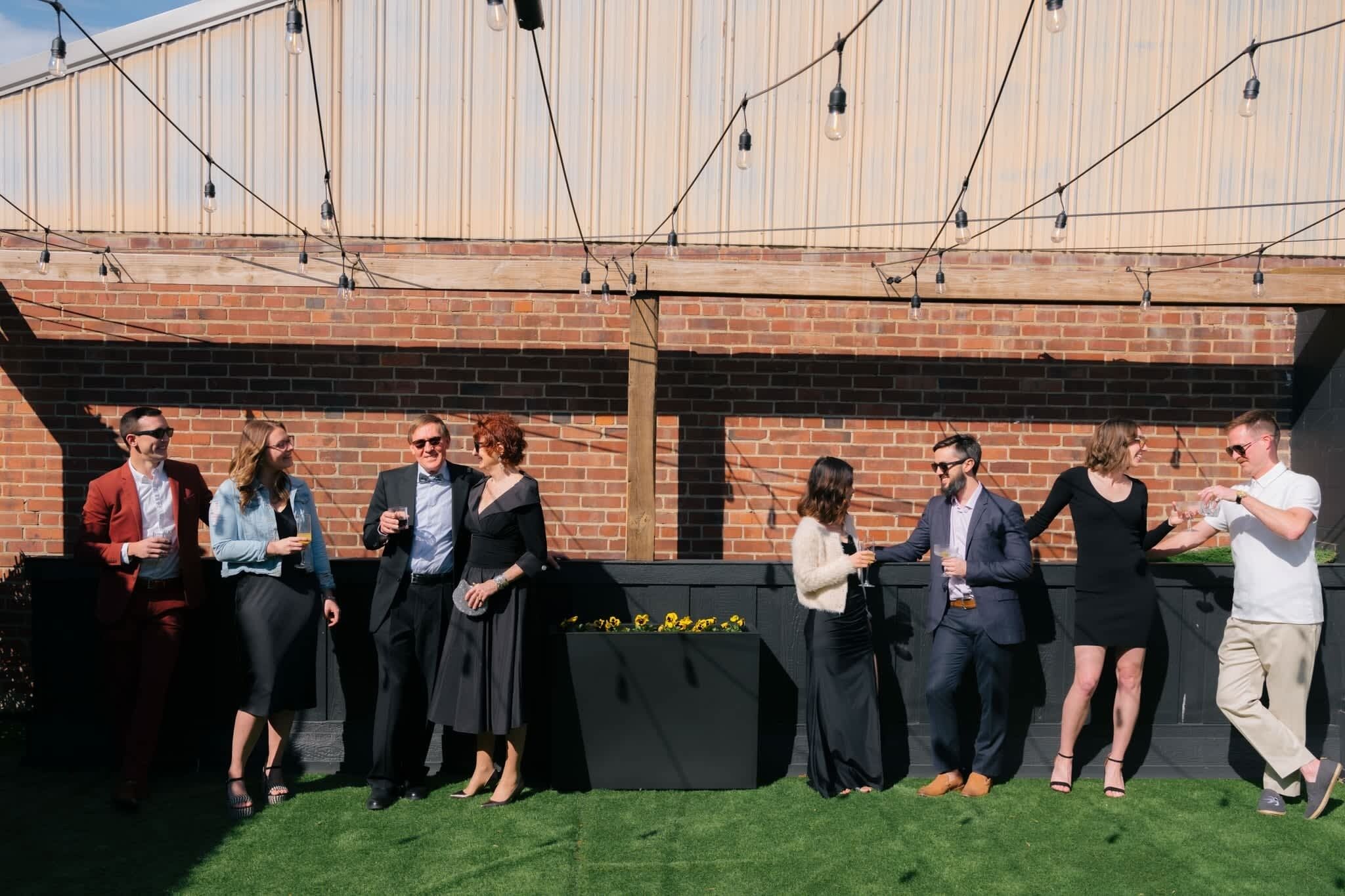 A group of people standing in a line outdoors, with a brick wall and string lights in the background.