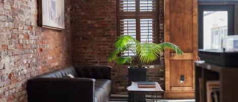 Een moderne woonkamer met een bakstenen muur, een zwarte leren bank, een groene fauteuil, een houten kast en een potplant.