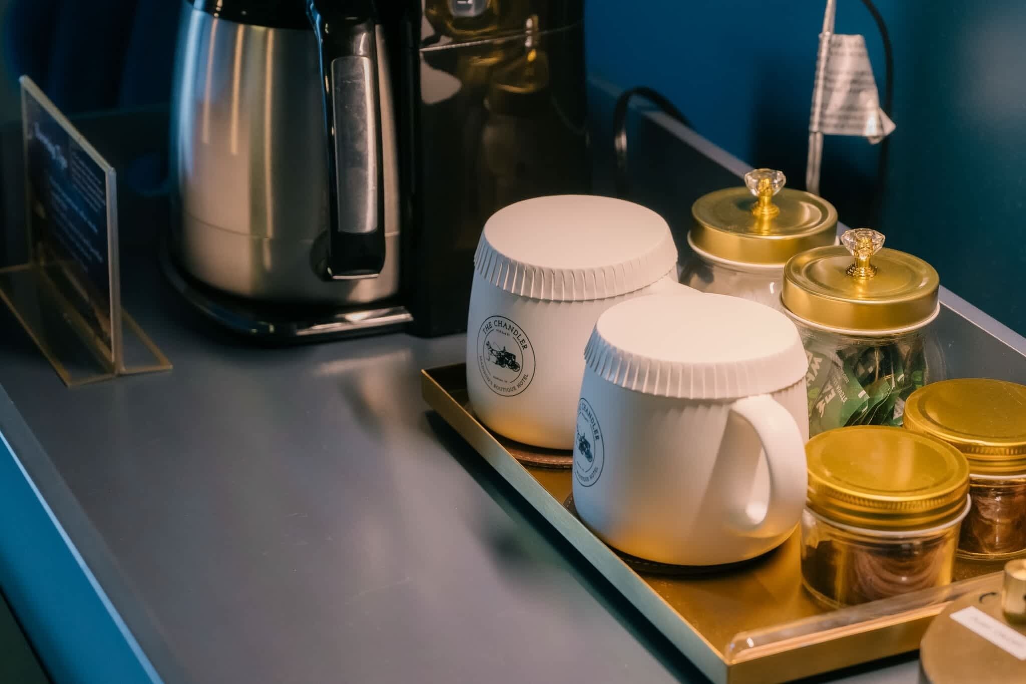 Una bandeja con tazas blancas y frascos dorados sobre una mesa, una cafetera y un hervidor.