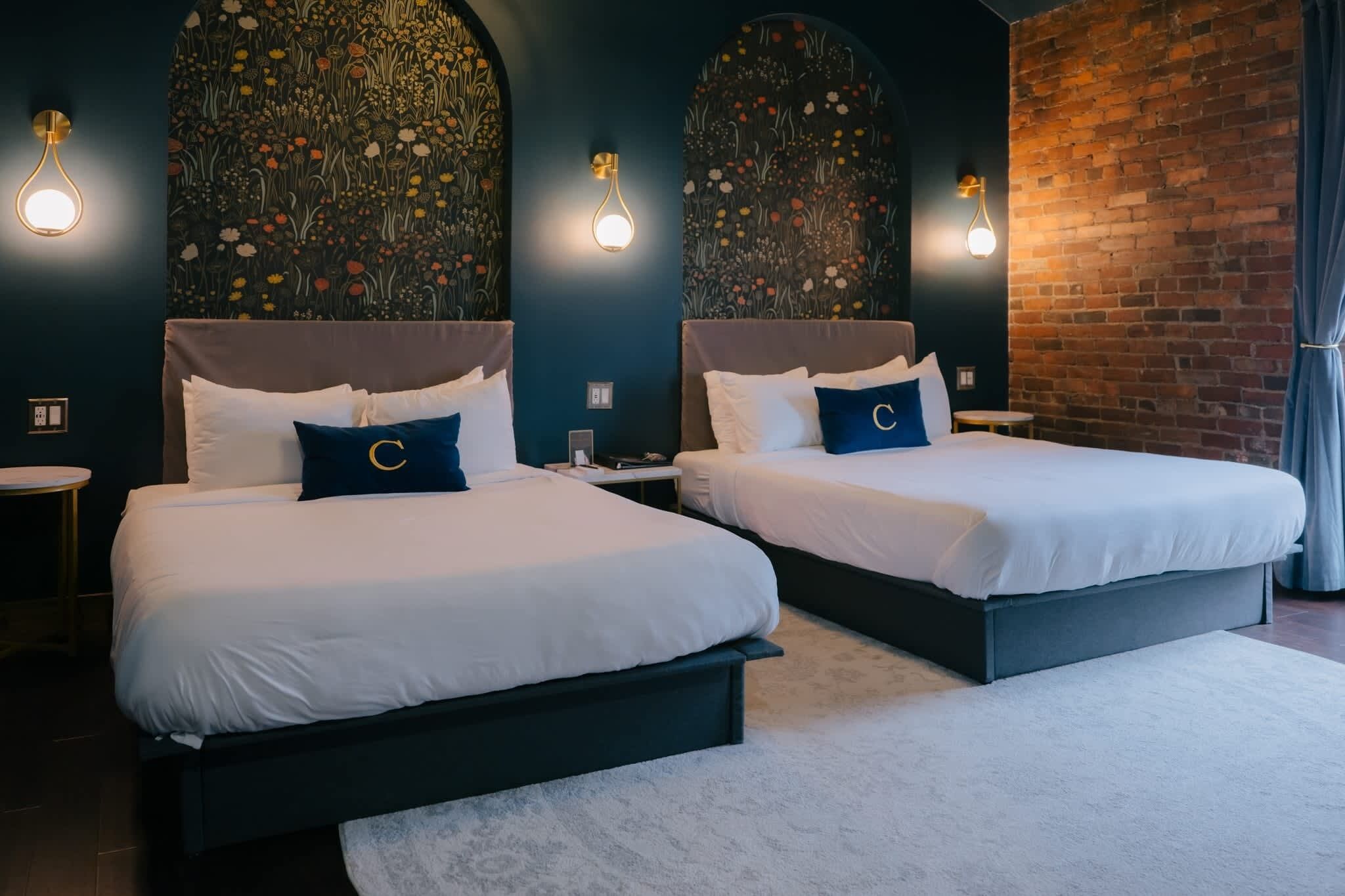 A hotel room with two beds, dark walls, floral wallpaper, and brick accents.