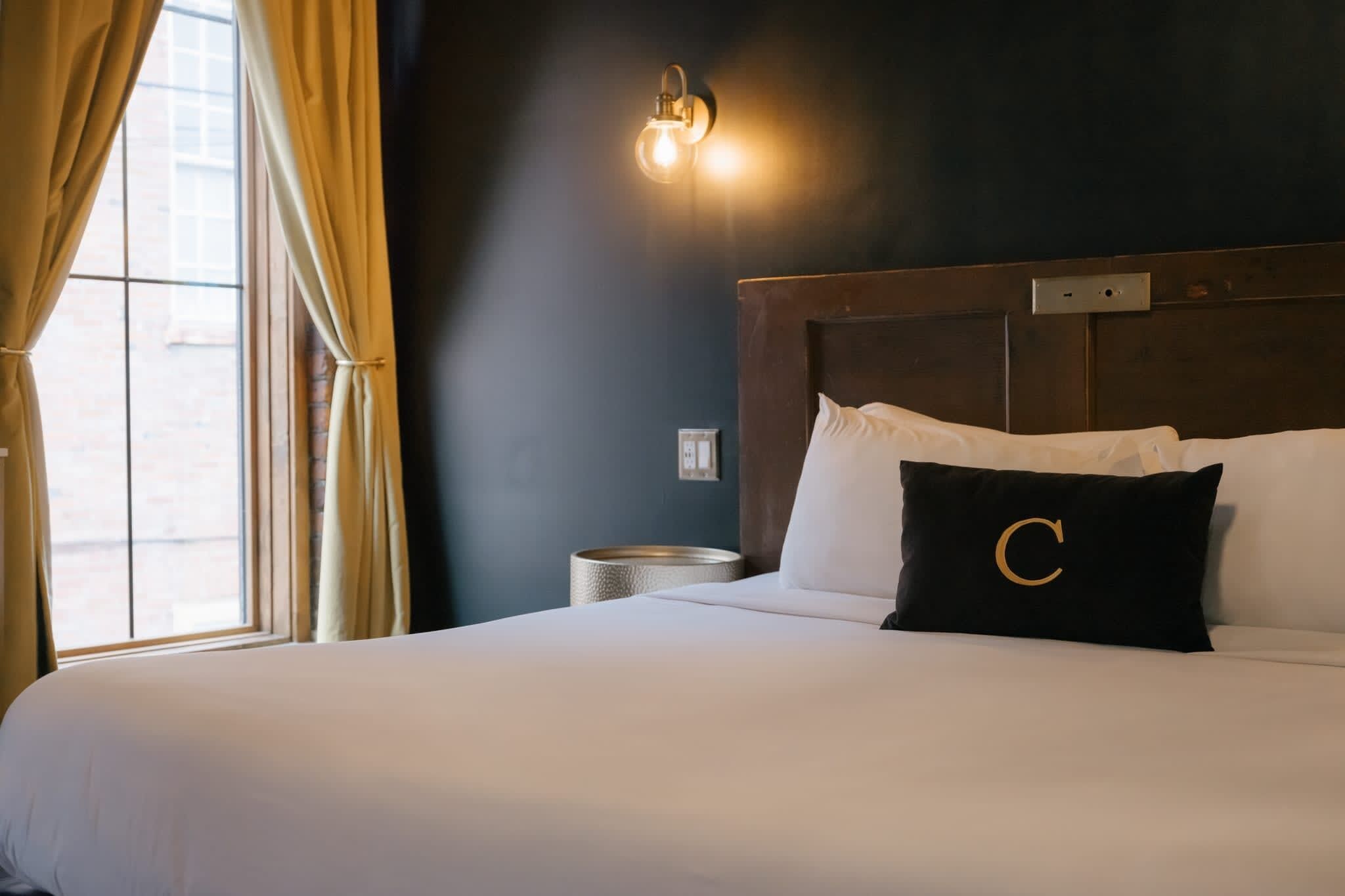 A hotel room with a large bed, a nightstand, a wall-mounted light, and a window with curtains.