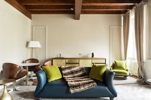 Una sala de estar con un sofá azul, almohadas verdes y marrones, una lámpara, un aparador de madera y una silla.