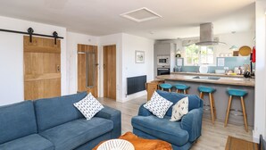 A modern living room with a sofa, armchair, and coffee table, a kitchen with a bar area, and wooden doors. - The Stables (Newport)