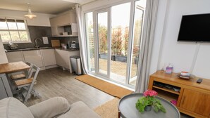 Una sala moderna con un sofá gris, una pequeña mesa redonda con una planta en maceta, una zona de cocina con un fregadero y armarios, y un televisor montado en la pared.