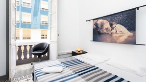 Habitación de hotel con cama, una silla, mesita de noche, un cuadro en la pared y ventana con vista al balcón.