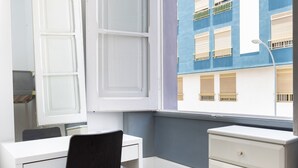 Una habitación con un escritorio blanco, una silla negra y un aparador blanco. Una ventana muestra un edificio azul con balcones.