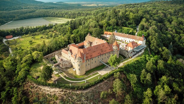 An aerial view of a castle with a large courtyard, surrounded by dense forest and rolling hills in the background.