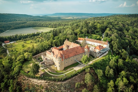 An aerial view of a castle with a large courtyard, surrounded by dense forest and rolling hills in the background.