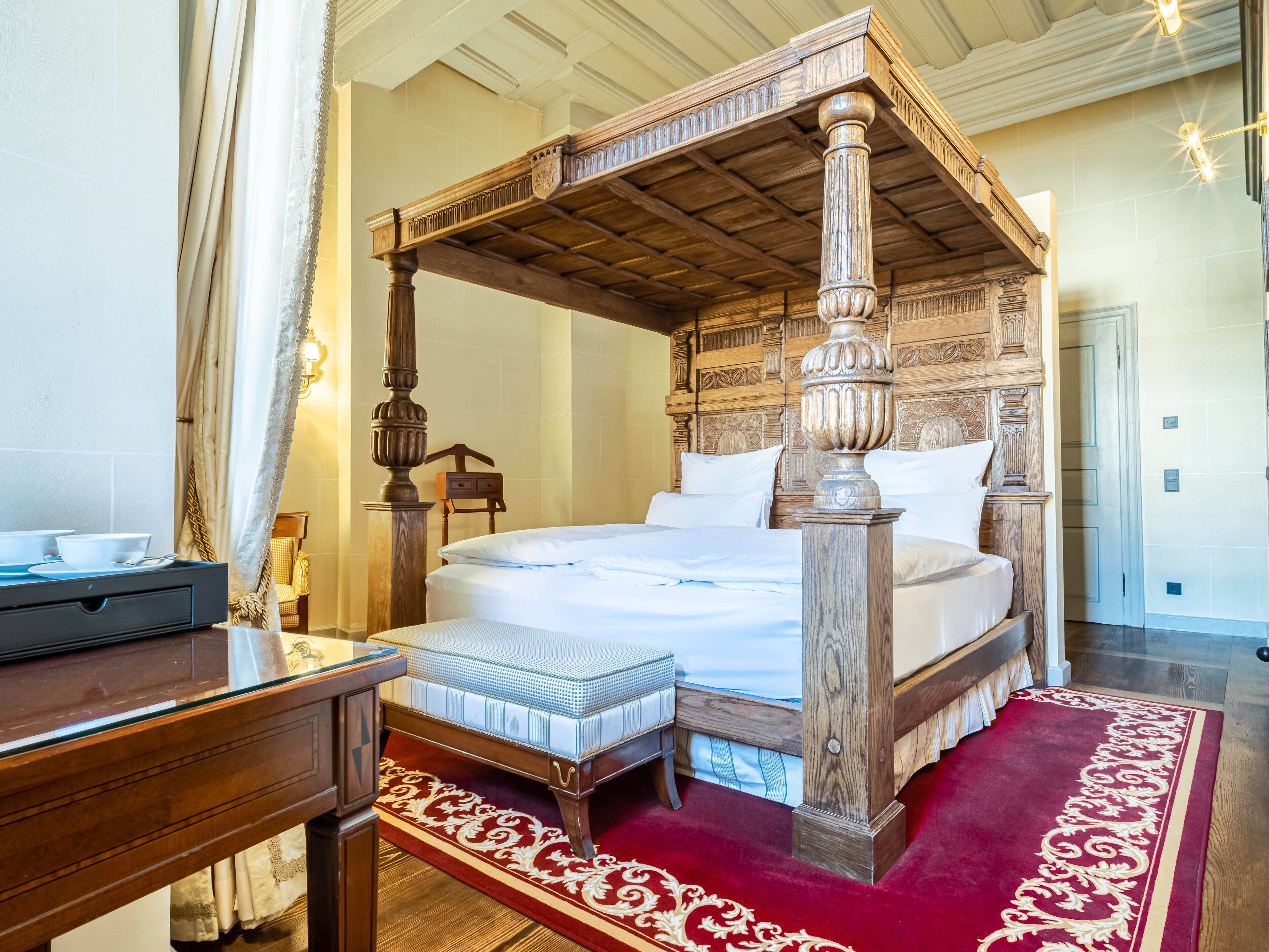 A four-poster bed with a wooden headboard, a red and white patterned rug, and a wooden nightstand with a tray.