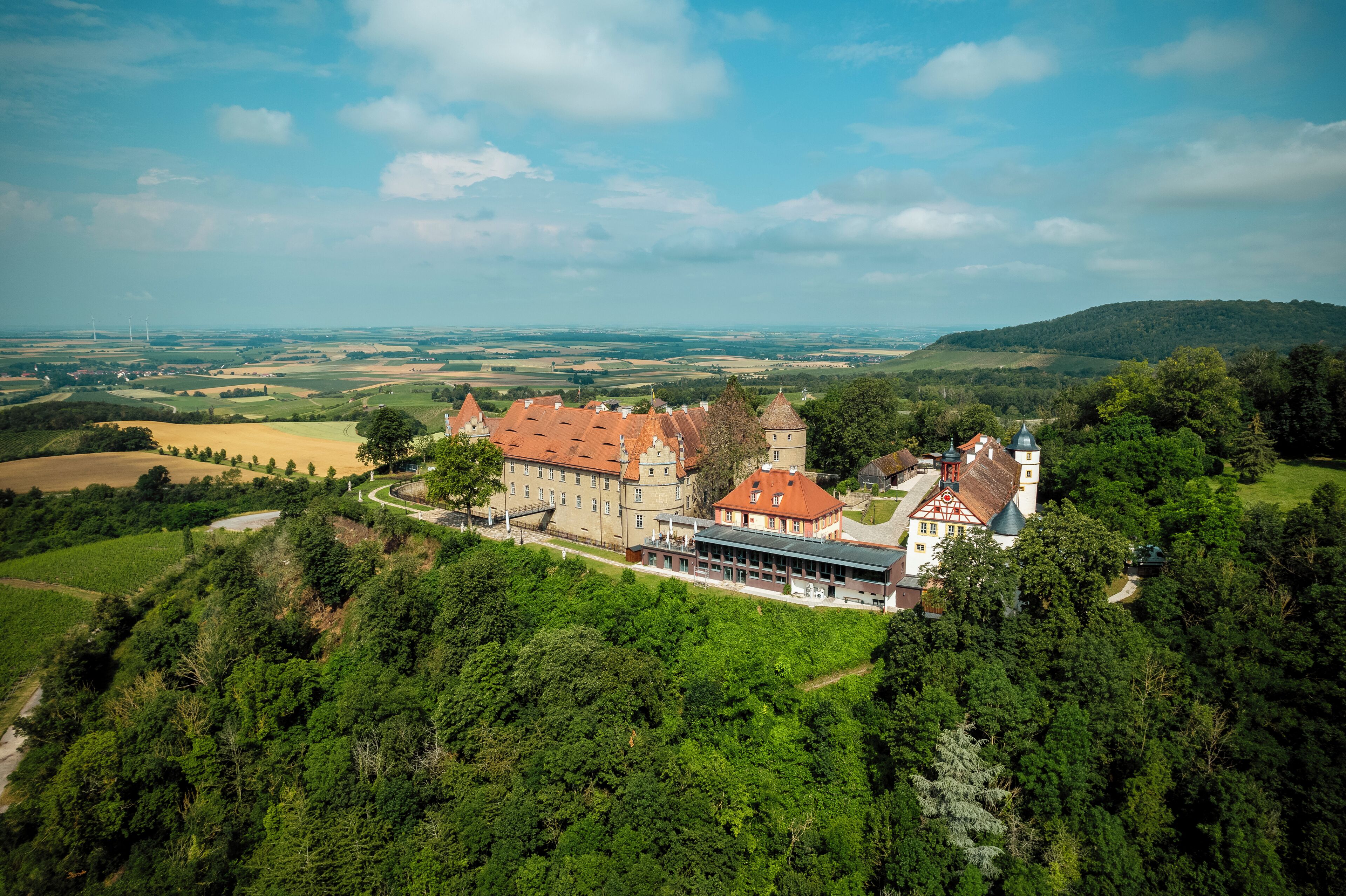 A large, historic building with multiple towers and a central courtyard, surrounded by lush greenery and fields.